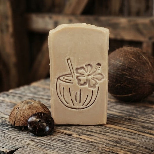 Black Coconut goat milk soap bar with engraved coconut drink design on rustic barnwood beside coconut shell pieces and a whole coconut.