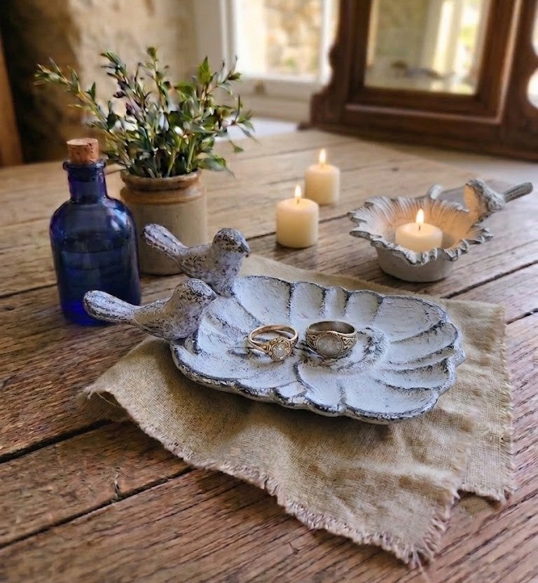 A rustic ceramic bird-shaped ring holder with a leaf-like base, styled on a burlap runner on a wooden table with candles, a blue glass bottle, and a small potted plant.