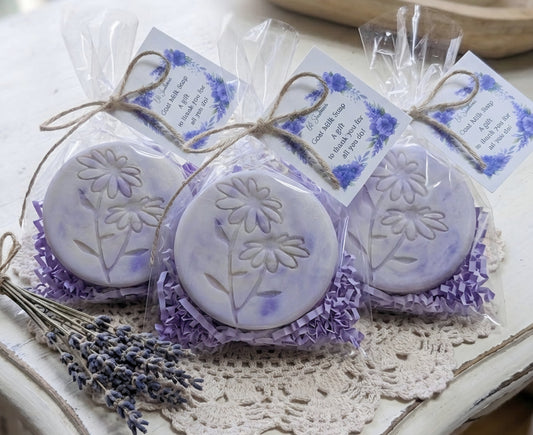 Three round lavender-scented soap bars with floral designs in clear packaging on a doily.