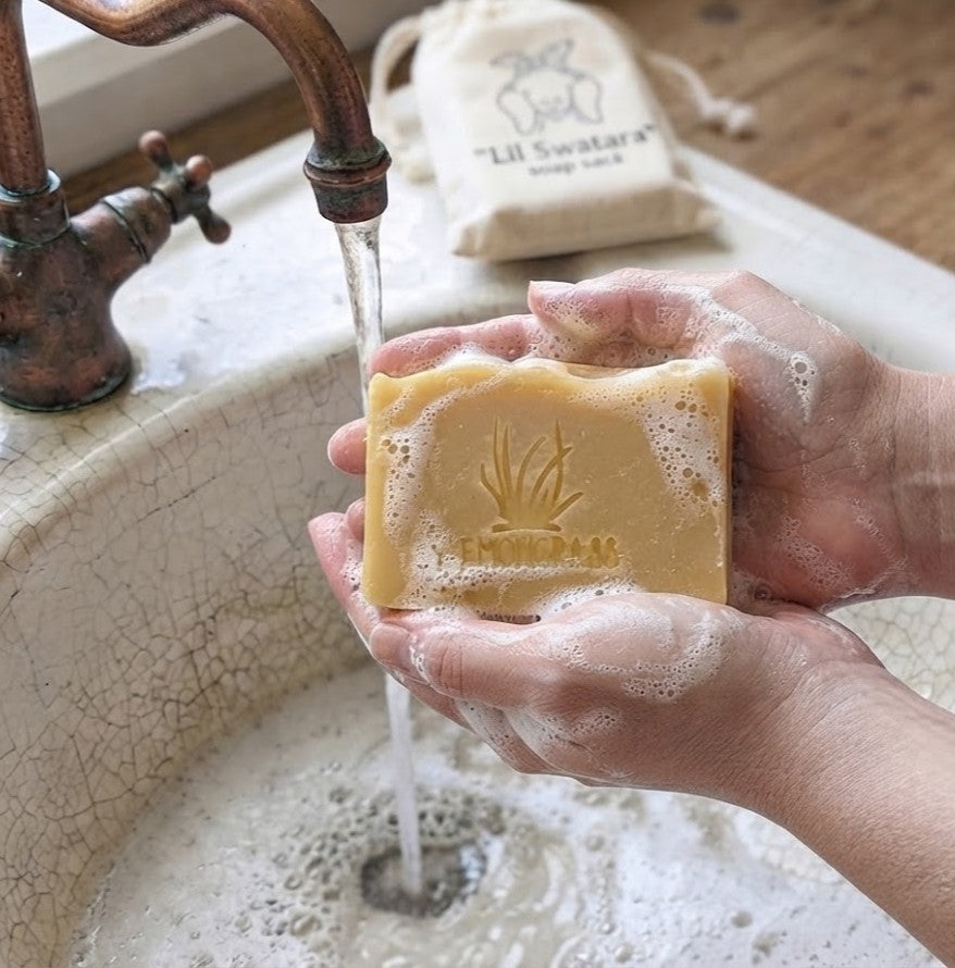Hands lathering a Natural Lemongrass goat milk tallow soap bar engraved with a lemongrass design under running water in a rustic sink.

