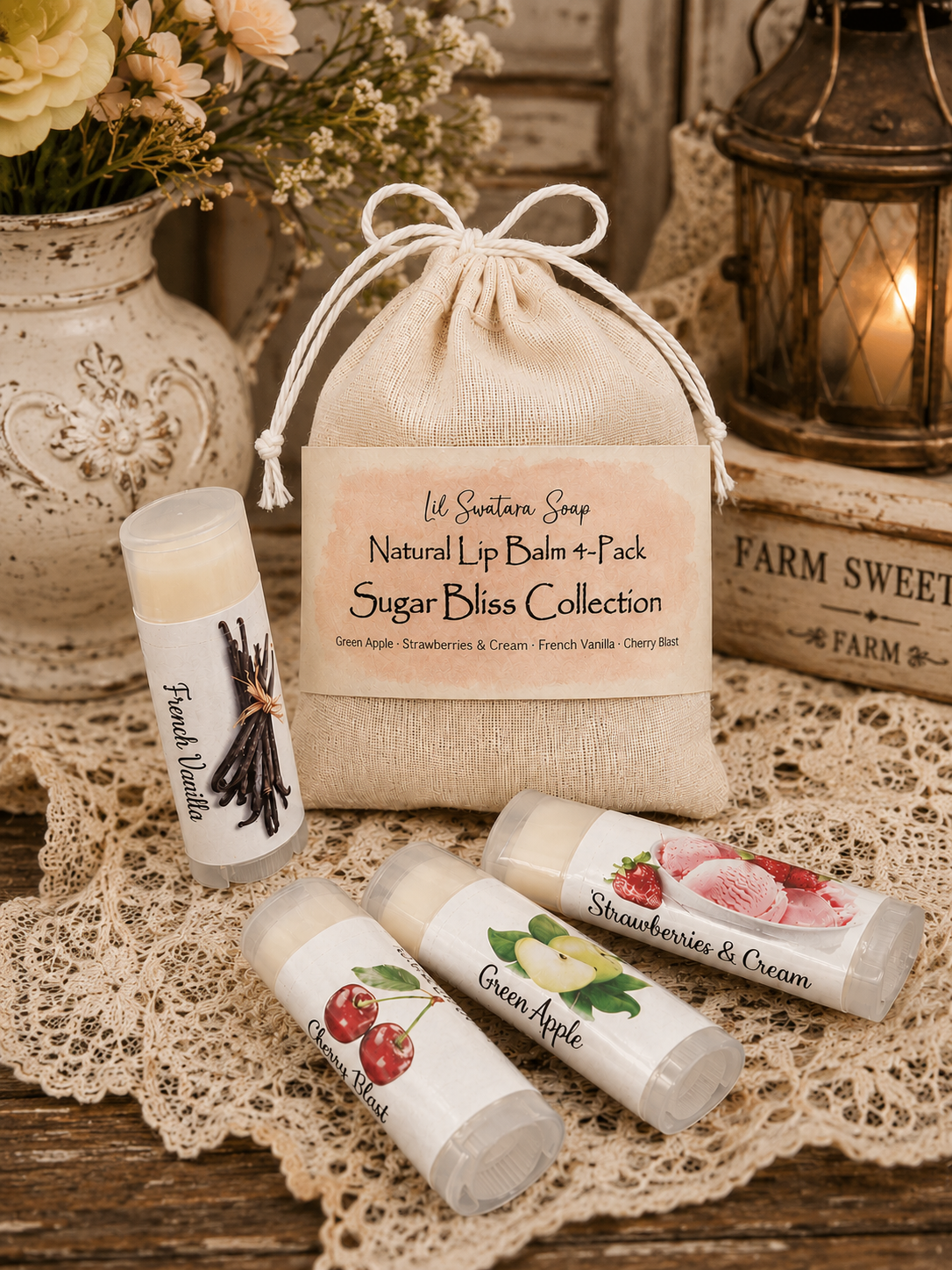 A natural lip balm and pack set from Ud Sanction Soap's Sugar Bliss Collection, featuring a beige burlap drawstring pouch with a printed label, alongside three clear tubes of lip balm with white labels showing illustrations of vanilla, green apple, and strawberries and cream. The products are styled on a rustic wooden table with a lace doily, a ceramic vase of dried flowers, and a lantern in the background.