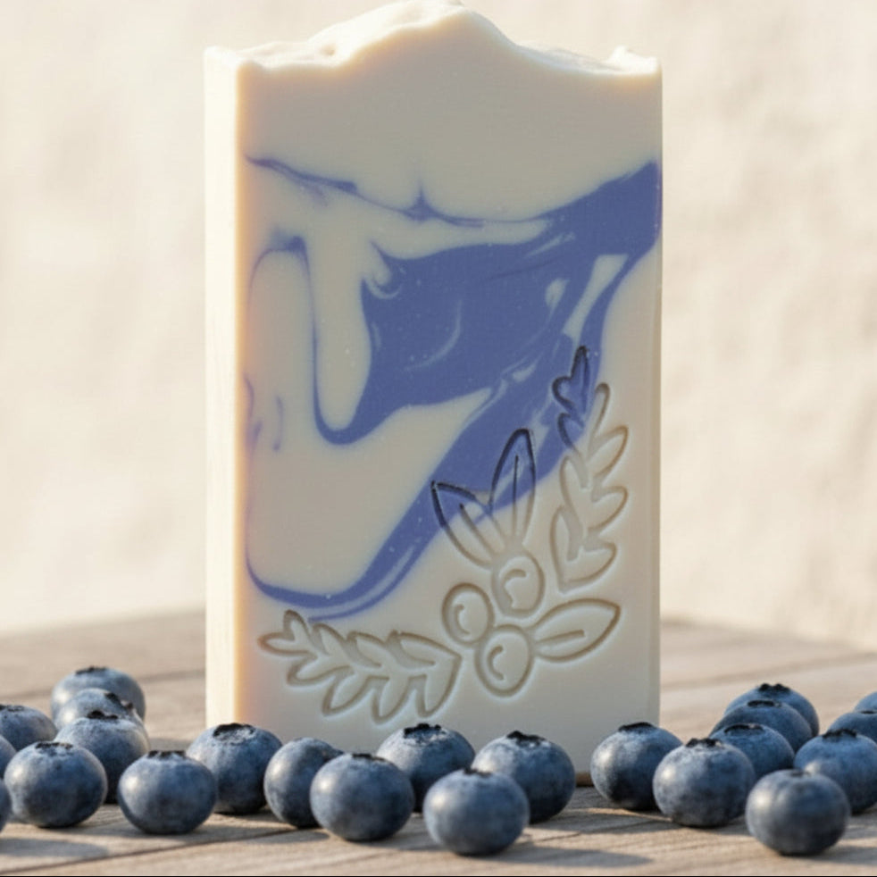 Handmade soap bars displayed outdoors on a wooden surface with blueberries and blackberries, alongside a small drawstring pouch. The soap has an off-white base with blue marbled swirls and embossed floral details.