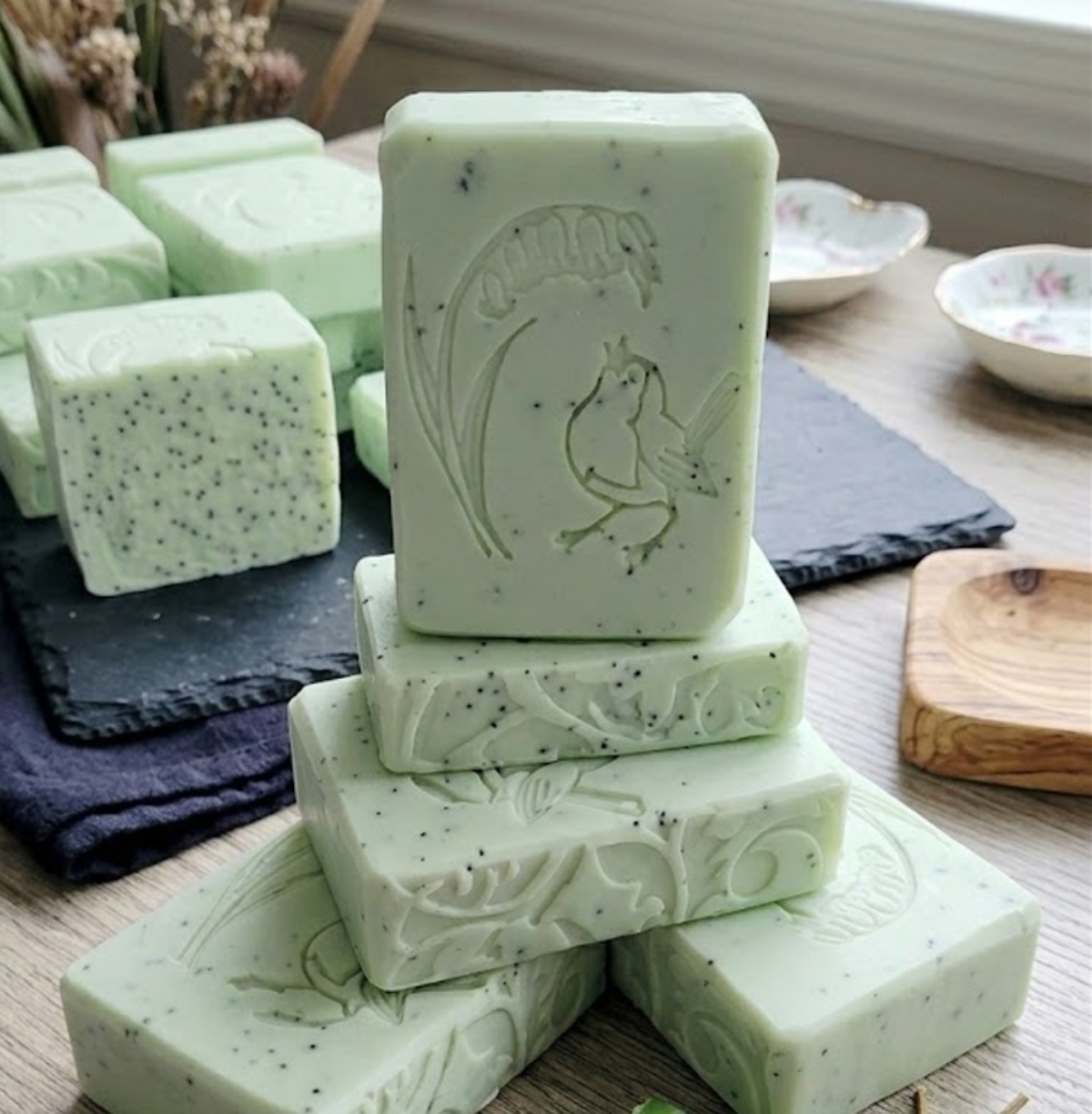 A stack of handmade pale green soap bars with black speckles and embossed floral and bird designs, displayed on a wooden surface with dark slate cloths and small bowls in the background.