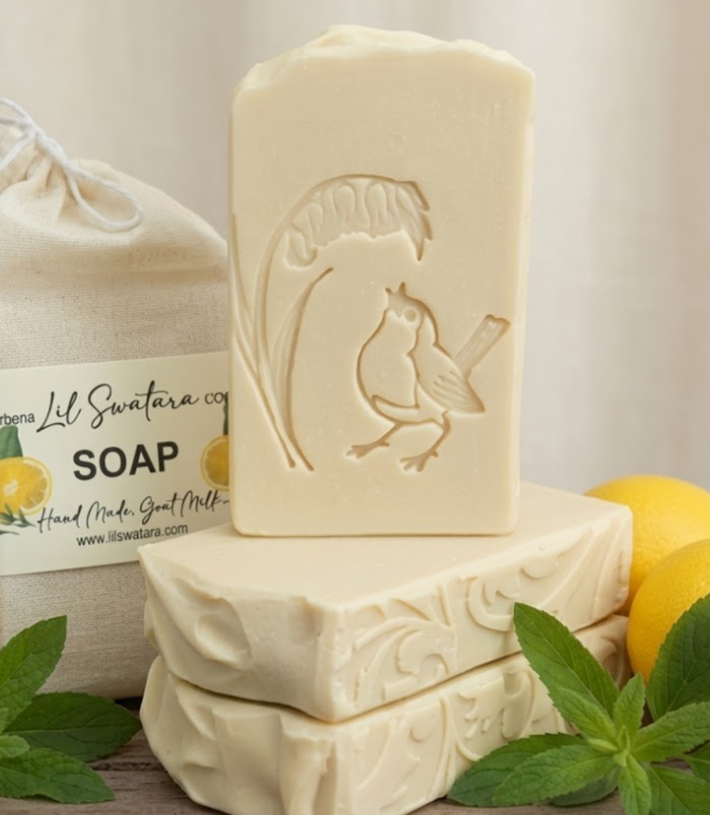 Three bars of handmade soap in an off-white/cream color, with one bar standing upright and embossed with a bird and leaf design. The soap bars are styled with fresh green mint leaves and whole lemons, with a drawstring bag of soap and a label in the background.