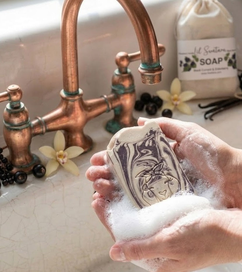 A close-up of a person's hands holding a bar of artisanal blackberry and magnolia scented goat milk soap and creating rich suds over a vintage white farmhouse sink. Fresh blackberries and a blooming magnolia flower are integrated into the lather, with an aged copper faucet and a fabric pouch of soap in the background.
