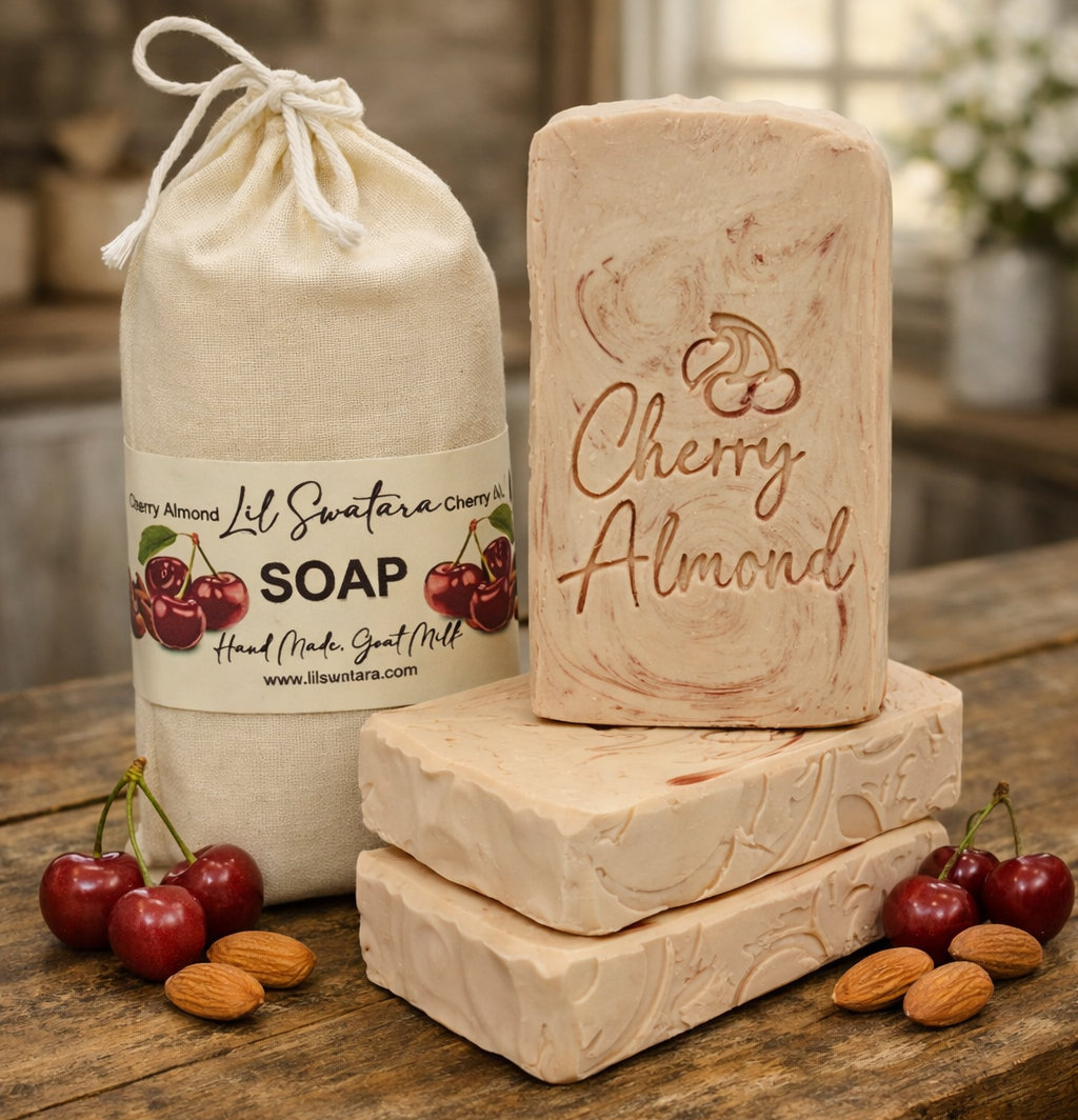 Cherry Almond Goat Milk Soap XL 6oz bar and matching drawstring sack displayed on a rustic farmhouse table with a few fresh cherries and almonds on each side. Creamy pink‑swirled handmade goat milk soap embossed with ‘Cherry Almond,’ highlighting its sweet, warm, and comforting scent.