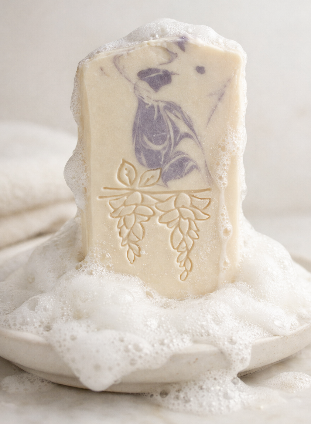 Handcrafted goat milk soap bar with creamy white lather and embossed botanical design on a ceramic dish.