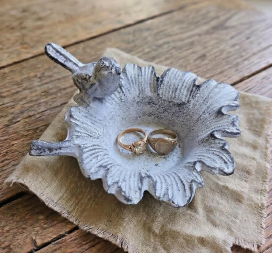 A rustic ceramic bird-shaped ring holder with a textured, distressed finish, resting on a piece of burlap fabric over a wooden table. Two gold rings are placed inside the holder.