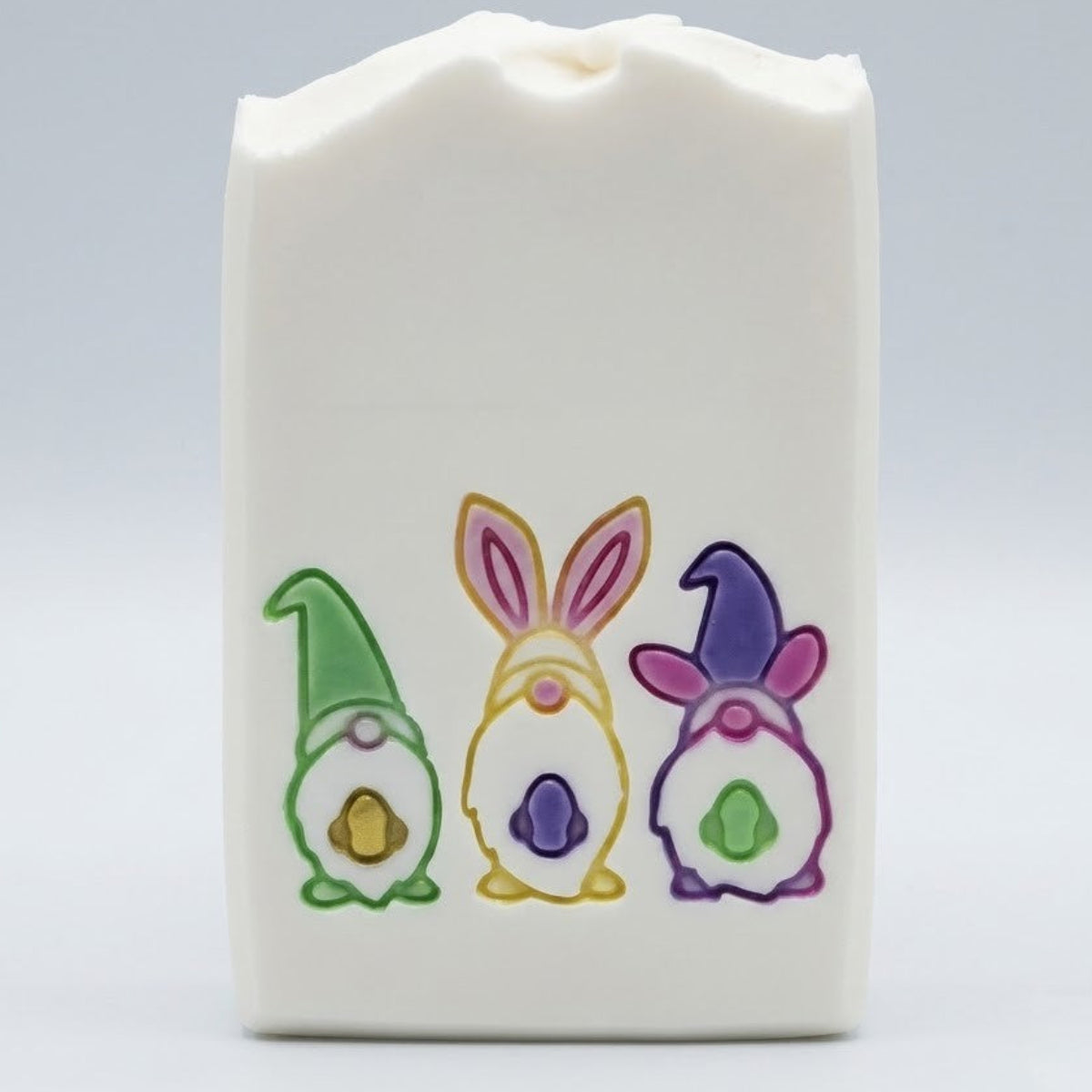 A white rectangular bar of soap with a wavy top edge, decorated with three colorful gnome and bunny illustrations in green, yellow, purple, and pink, set against a light gray background.