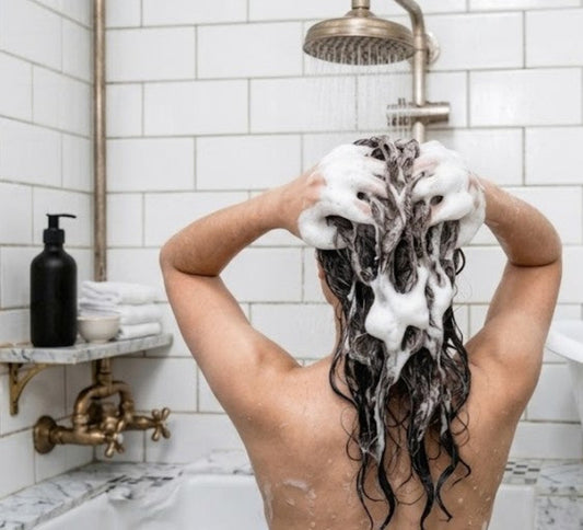 Person washing hair under shower with thick creamy goat milk shampoo lather, bright clean bathroom scene.