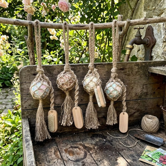Four handmade jute‑wrapped garden soap balls with tassels hanging from a branch above a rustic garden bench with tools and seed packets.