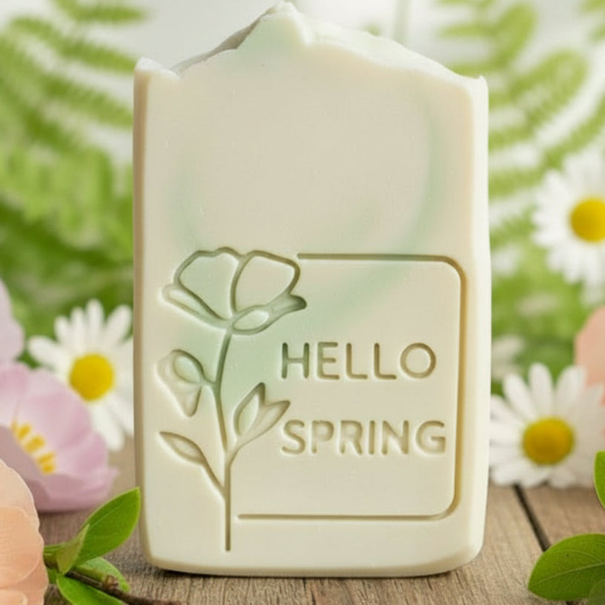A rectangular bar of cream-colored soap with a floral embossed design and the text 'HELLO SPRING' on the front, displayed on a wooden surface surrounded by pastel flowers.