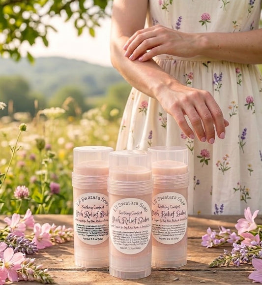 Woman in a soft floral dress applying itch relief balm to her forearm outdoors, with Lil Swatara Soap Itch Relief Balm sticks on a wooden table surrounded by wildflowers.