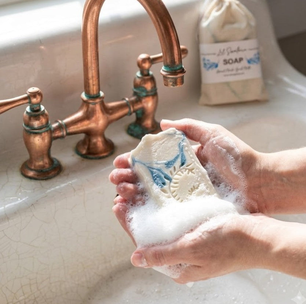 Hands lathering a bar of handmade Lil Swatara goat milk soap with a creamy lather in a vintage copper bathroom sink.