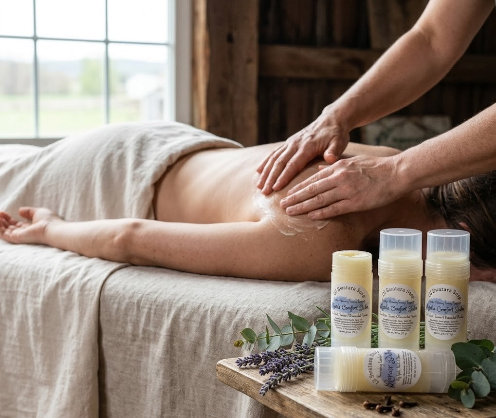 Muscle Comfort Balm displayed on a wooden table with lavender and greenery beside a towel‑covered back receiving soothing muscle relief in a rustic room.