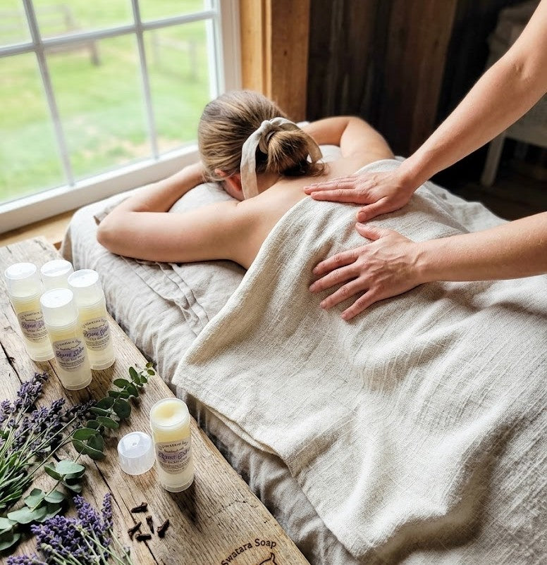 Towel‑covered back receiving muscle relief while Muscle Comfort Balm sits on a rustic wooden table with lavender and greenery in soft farmhouse light.