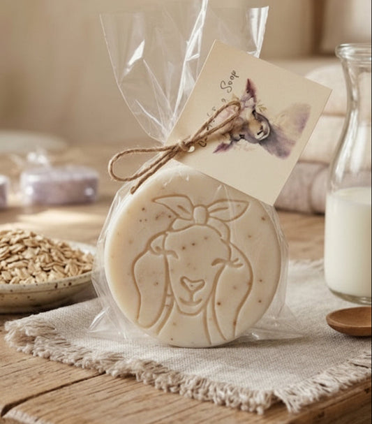 A creamy, natural goat milk soap puck infused with soothing oats, rich milk, and sweet honey for a gentle, moisturizing cleanse. This circular soap has a detailed engraving of a goat's face and is beautifully presented in a clear gift bag with a rustic twine bow.