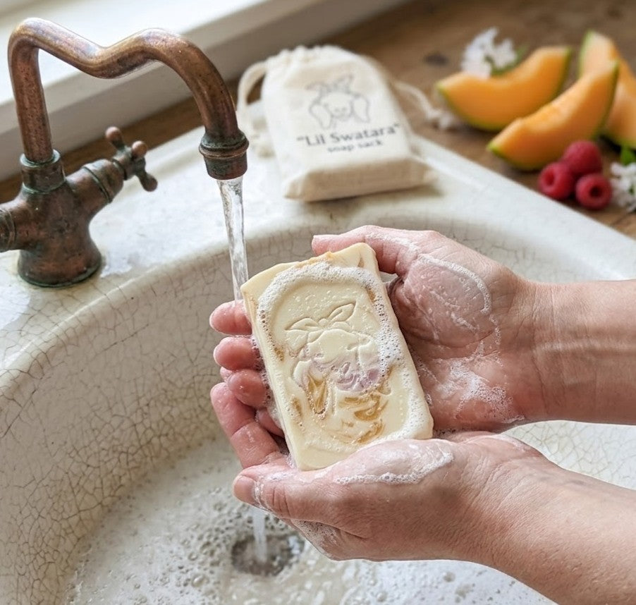 Hands cupping a creamy, embossed Raspberry Melon goat milk soap bar under a vintage copper faucet in a porcelain basin, with a Lil Swatara branded muslin soap sack and fresh melon and raspberry garnishes in the background.