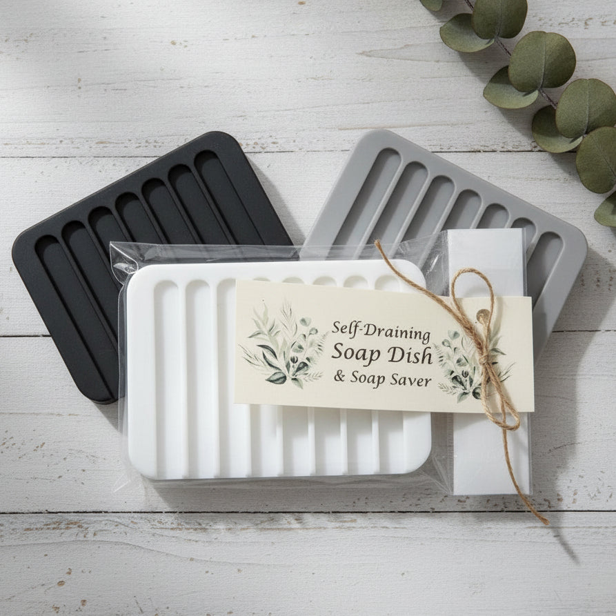 A white self-draining soap dish and soap saver set wrapped in clear plastic, with a cream paper tag tied with twine that reads 'Self-Draining Soap Dish & Soap Saver.' The set is photographed on a white wood surface with eucalyptus leaves in the corner.