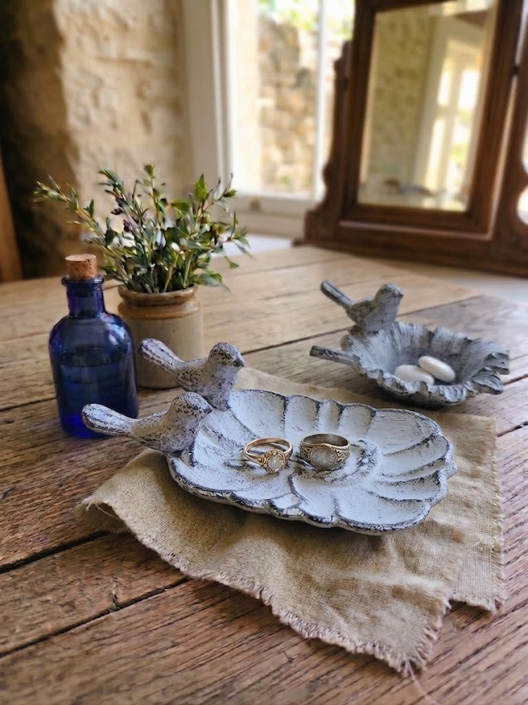 Three rustic bird-shaped ring holders made of distressed gray stone or ceramic, displayed on a weathered wooden table with a burlap cloth, a blue glass bottle, and a small potted plant.