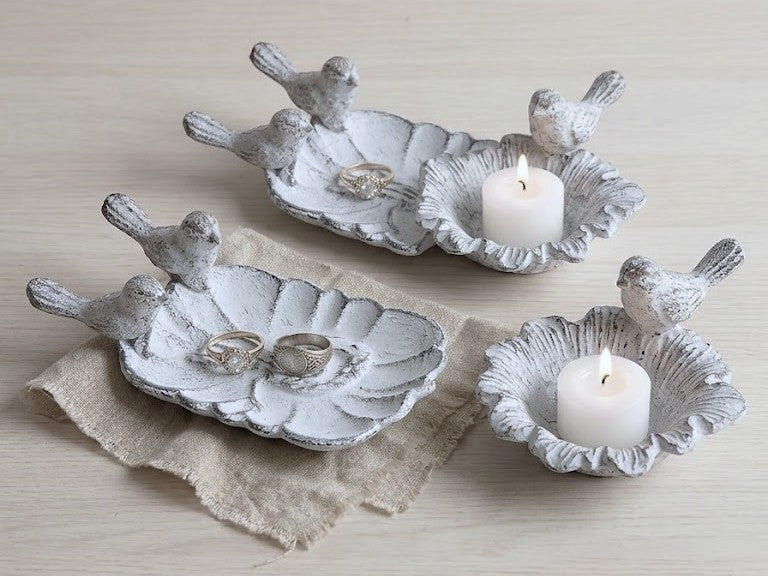 Three white ceramic bird-shaped dish holders with a distressed finish, arranged on a light wood surface. Two hold rings, and one holds a lit white pillar candle.