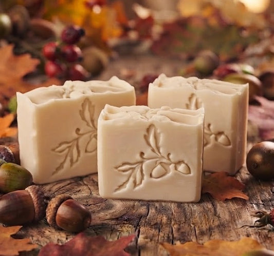 Three rectangular bars of handmade soap showing acorn soap stamp with embossed leaf and acorn designs, displayed on a rustic wooden surface surrounded by autumn leaves, acorns, and berries.