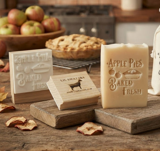 Soap stamp image of A styled product display featuring a cream-colored soap bar and wooden blocks stamped with 'Apple Pies Baked Fresh,' alongside a cream drawstring pouch with matching text, arranged on a rustic wooden table with apples, cinnamon, and a pie in the background.