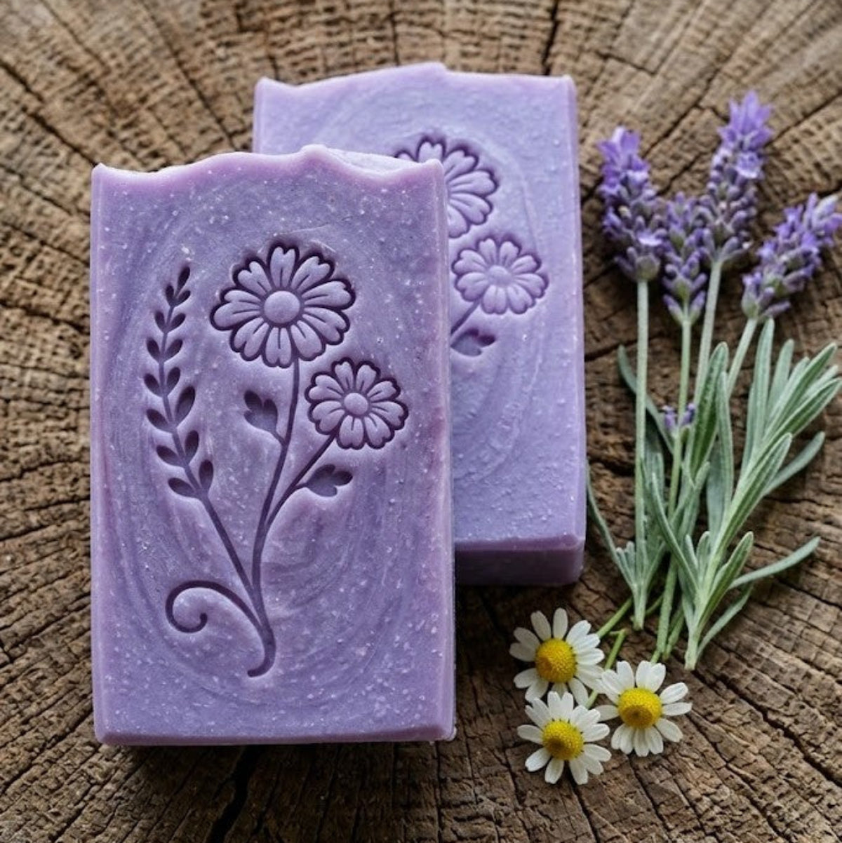 Two lavender-colored handmade soap bars with soap impression from stamp in lavender and chamomile and leaf patterns, displayed on a rustic wood slice background alongside fresh lavender stems and small white daisies.