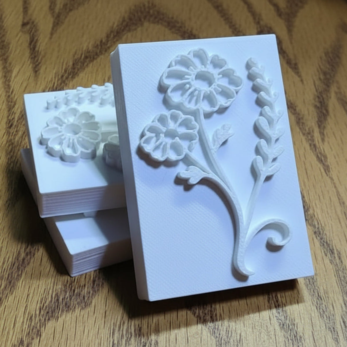 A white rectangular floral soap stamp or embosser with raised flower and leaf designs, shown standing upright on a wood-grain surface with a stack of similar stamps behind it.