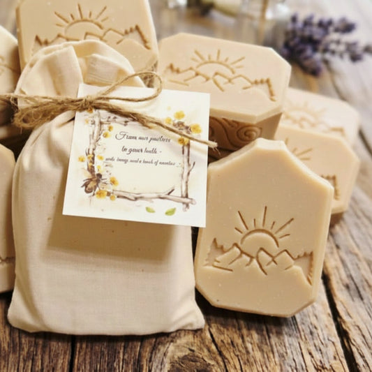 Handmade natural soap bars with soap stamped mountain and sun designs, arranged around a small beige fabric gift bag tied with twine and a decorative tag, set on a rustic wooden surface.
