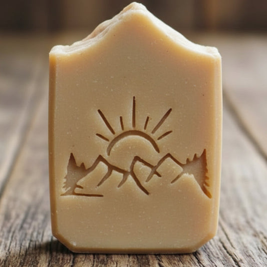  handmade soap bars, placed on a rustic wooden surface. The soaps are light tan with embossed nature-themed designs mountains, trees, and sun