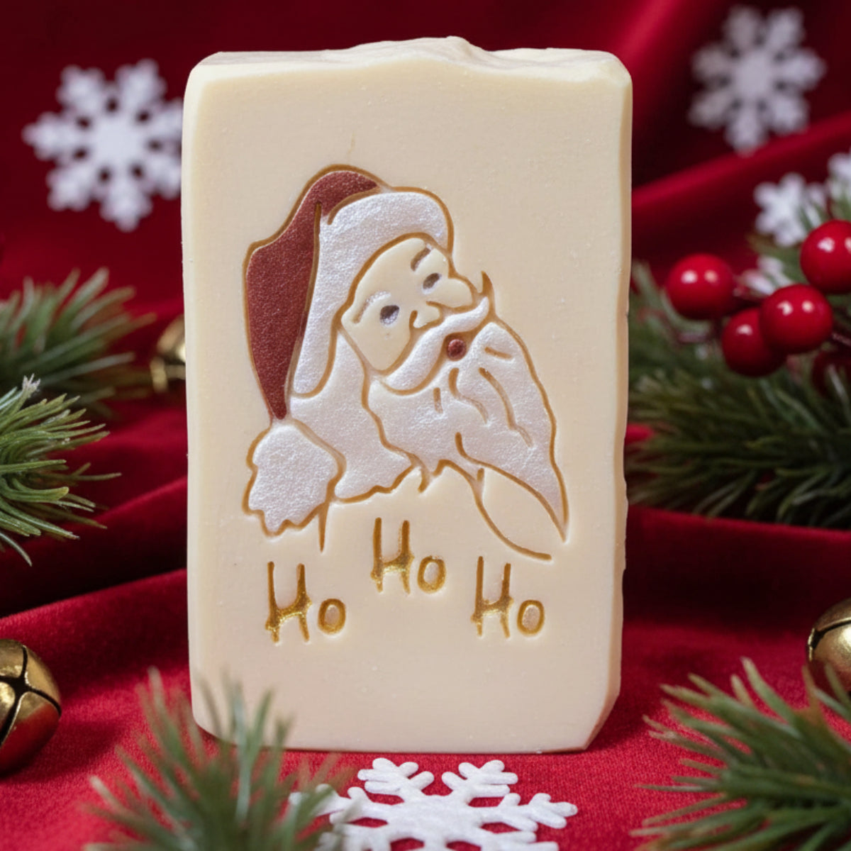 A close-up of handmade rectangular soap bars decorated with a Santa Claus face and the words 'Ho Ho Ho' in gold, with more similar soaps arranged in the background.