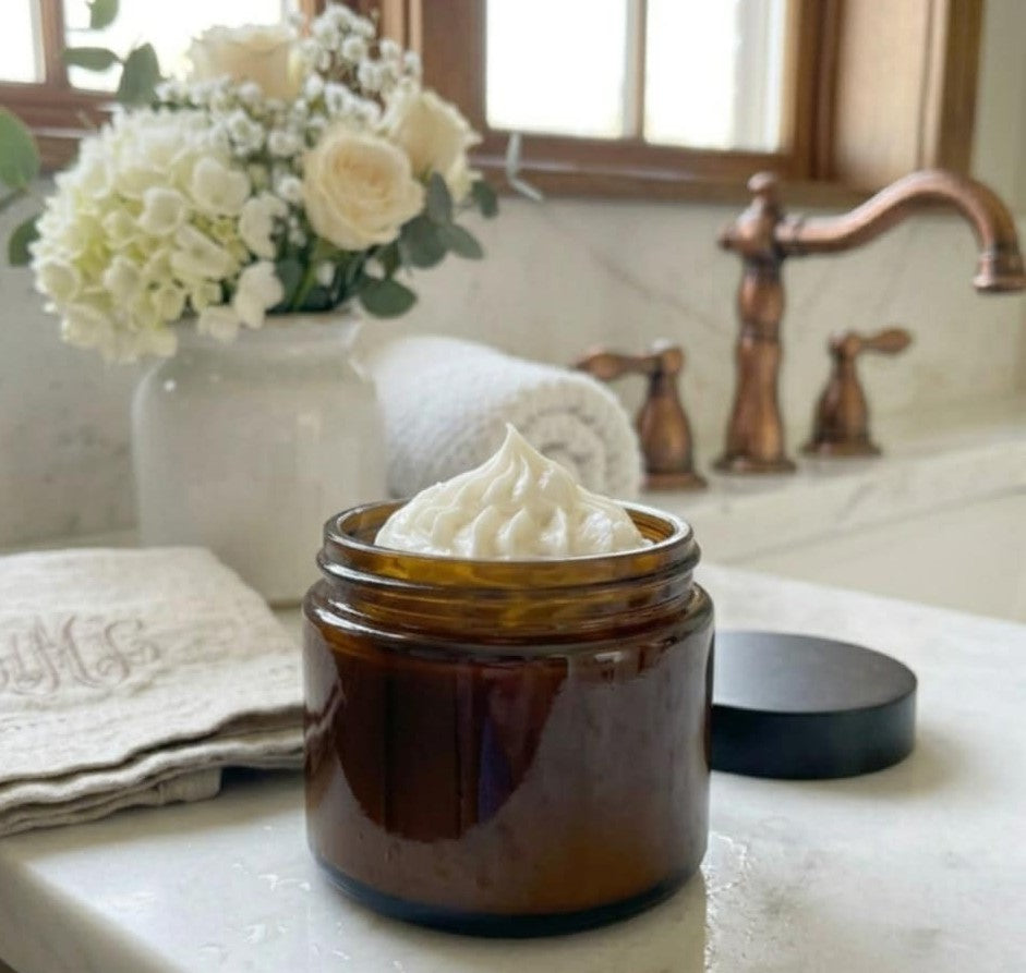 Whipped tallow balm in a brown glass jar on a marble bathroom counter with white flowers and towels, luxury natural skincare scene.