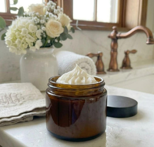 Whipped tallow balm in a brown glass jar on a marble bathroom counter with white flowers and towels, luxury natural skincare scene.