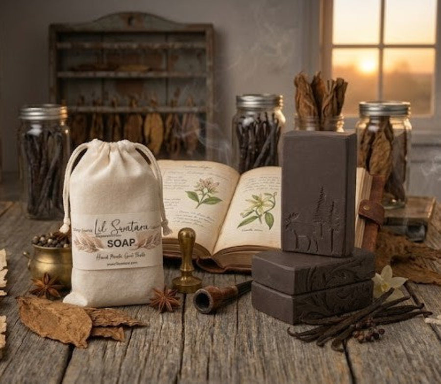 Handcrafted Tobacco Vanilla goat milk soap bars stacked beside a muslin drawstring bag labeled Lil Swatara Soap, surrounded by dried tobacco leaves, vanilla beans, and cream‑colored vanilla flowers on a rustic wooden surface.