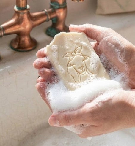 vintage white farmhouse-style sink where a pair of hands is washing with a bar of goat milk soap. The large, weathered copper kitchen faucet with two handles and patina stands prominently. The cream-colored soap bar has an engraved illustration of a goat with a bandana on its head and is covered in white soap lather and foam in the hands. In the background to the right, a reusable cloth drawstring bag labeled 'Lil Swatara SOAP'