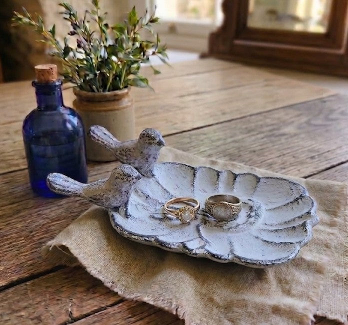 A rustic ceramic ring tray shaped like a leaf with two small bird figures on the left side, holding two rings, displayed on a burlap cloth on a wooden table with a blue glass bottle and potted plant in the background.